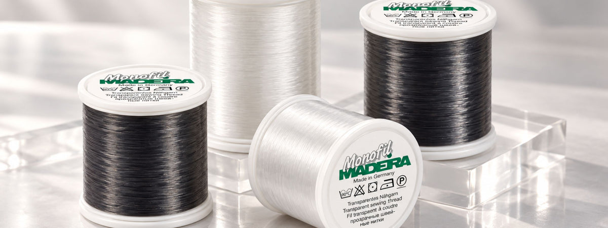 Four Madeira Monofil transparent sewing thread spools in clear and smoke colors, arranged on a reflective marble surface with clear acrylic risers and soft natural lighting.