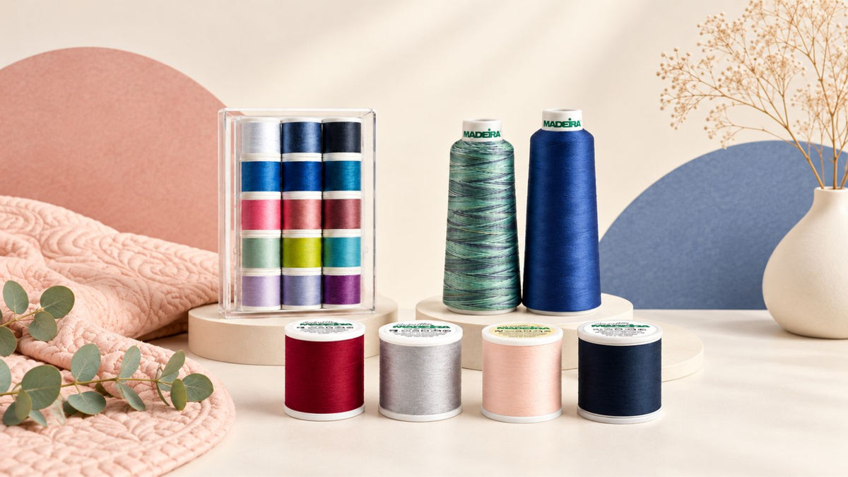 Assortment of Madeira quilting and sewing threads featuring colorful thread spools, blue and variegated thread cones, and a thread collection box displayed alongside a soft pink quilt and decorative greenery.